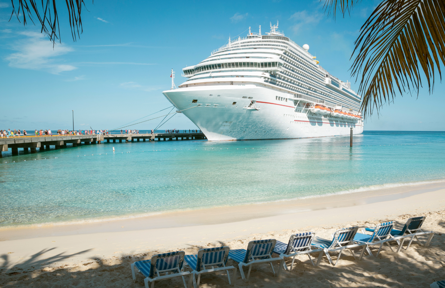 Large white cruise ship docked at a tropical port in the Caribbean, viewed from a sandy beach with lounge chairs and turquoise water.